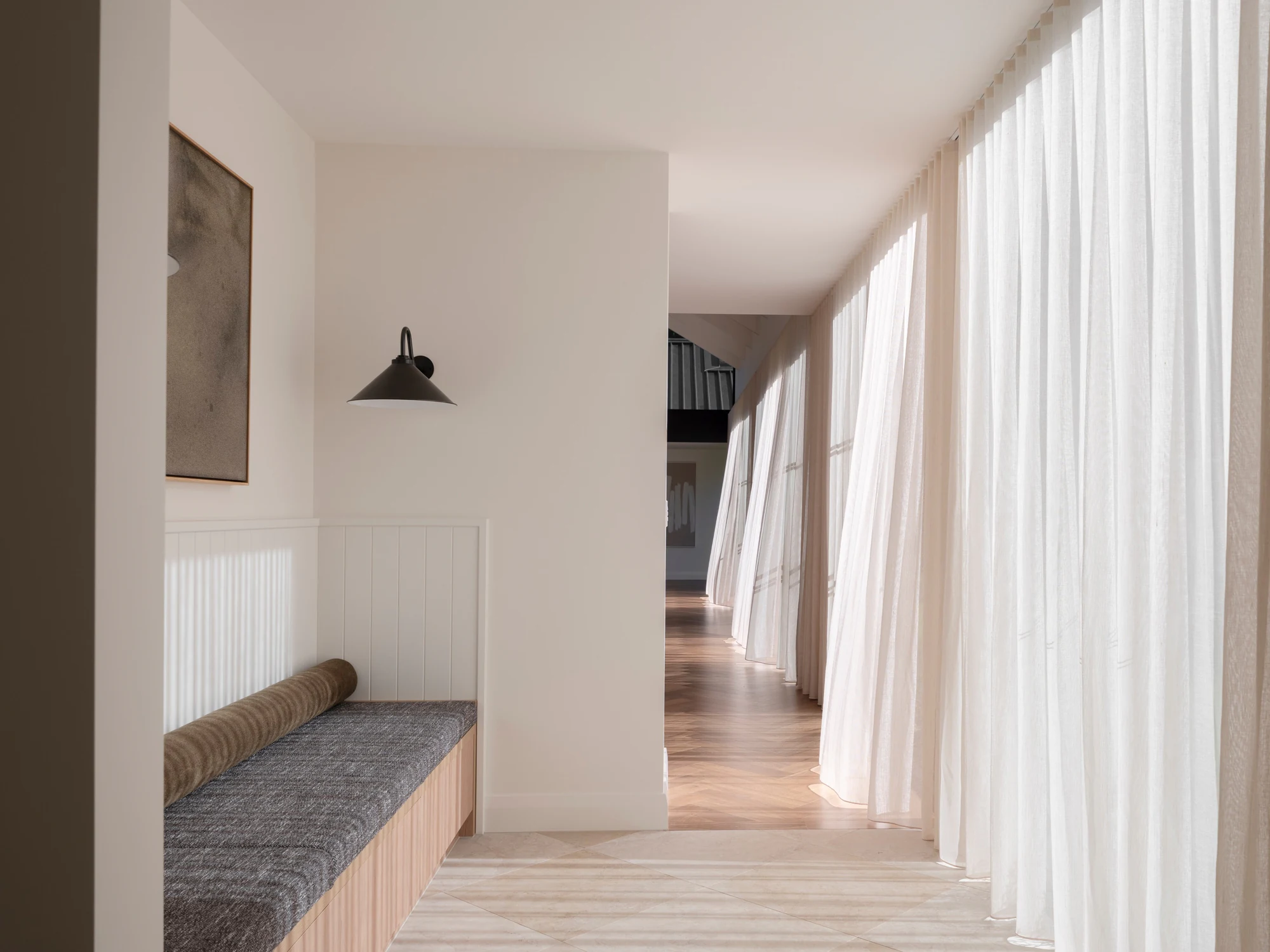A built-in bench seat and black wall light beside a sunlit hallway with sheer curtains.